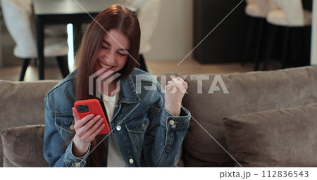 Young woman rejoices at the message on smartphone. Feel amazed happy and saying WOW on home background. Big win good news celebrate lottery jackpot doing winner gesture. 112836543