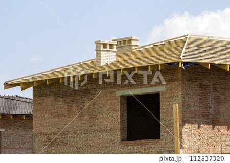 Close-up detail of wooden roof of new brick house with two white chimneys under construction. Timber frame of natural materials against bright sky. Professional building and reconstruction concept. 112837320