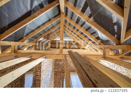 Attic of a building under construction with wooden beams of a roof structure and brick walls. Attic of a building under construction with wooden beams of a roof structure and brick walls. 112837395
