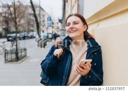 Happy cheerful young woman with backpack walking on city street checks her smartphone. Portrait of beautiful 30s girl using smartphone outdoors. Urban lifestyle concept. Happy cheerful young woman with backpack walking on city street checks her smartphone. Portrait of beautiful 30s girl using smartphone outdoors. Urban lifestyle concept. 112837957