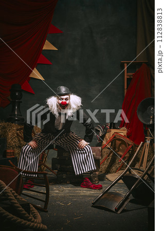 Clown in black hat, white face with red nose and striped pants playing over dark retro circus backstage background. Comedian 112838813
