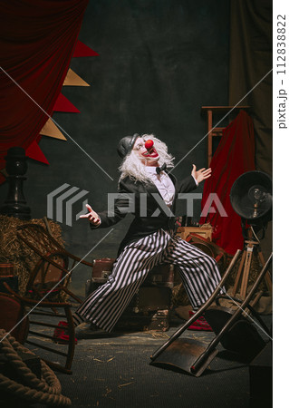 Clown emotional man in black hat, white face with red nose and striped pants playing over dark retro circus backstage background. 112838822