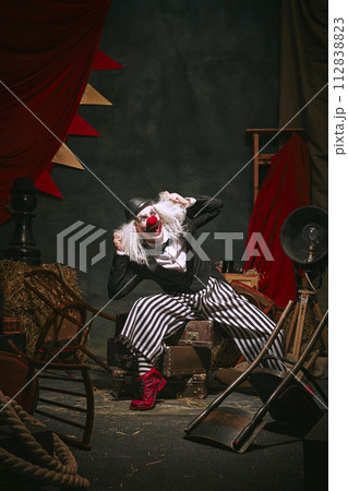 Clown in black hat, white face with red nose and striped pants shouting over dark retro circus backstage background. 112838823