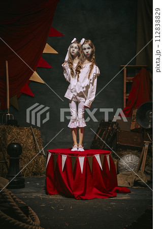 Siamese twin girls in white vintage costumes with makeup stand on stage over dark retro circus backstage background. Spooky-themed magic show 112838829