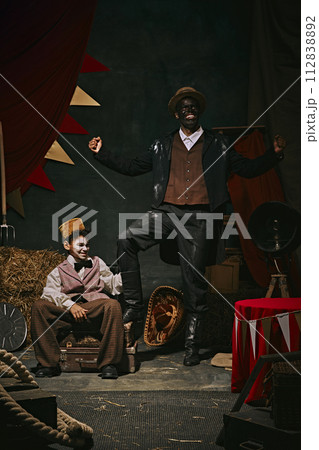 High African man with dirty face standing with little servant boy over dark retro circus backstage background. 20th-century vaudeville era. High African man with dirty face standing with little servant boy over dark retro circus backstage background. 20th-century vaudeville era. 112838892