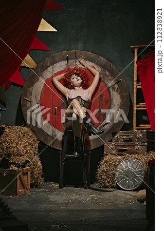 Danger. Elegant young woman with bright makeup and stage costume sitting next to target with knives over dark retro circus backstage background. 112838921