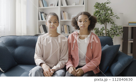 Two African American sisters sitting on sofa and seriously looking at camera Two African American sisters sitting on sofa and seriously looking at camera 112840630