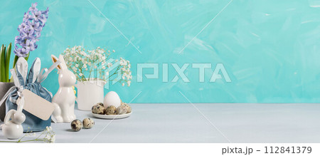 Easter banner. Eggs, ceramic bunnies, flowers, gift bag, blank note on turquoise. Copy space. Easter banner. Eggs, ceramic bunnies, flowers, gift bag, blank note on turquoise. Copy space. 112841379