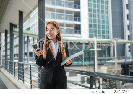 A young Asian businesswomen wearing a suit holding files standing in a big city on a busy downtown street. Young Asian businesswoman using smartphone texting to contact clients. 112842270