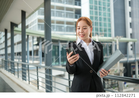 A young Asian businesswomen wearing a suit holding files standing in a big city on a busy downtown street. Young Asian businesswoman using smartphone texting to contact clients. 112842271
