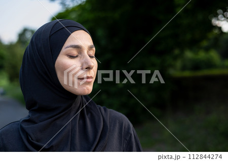 A Muslim woman wearing a hijab stands amidst lush greenery, symbolizing serenity and the connection between faith and nature. 112844274