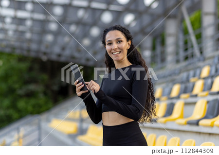 A smiling young woman wearing athletic wear holds a smartphone while standing at a sports stadium with seats in the background. 112844309