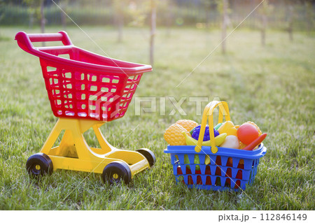 Shopping pushcart and a basket with toy fruits and vegetables. Bright plastic colorful toys for children outdoors on sunny summer day. 112846149