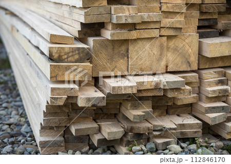 Close-up of piled stack of natural brown uneven rough wooden boards lit by bright sun. Industrial timber for carpentry, building, repairing and furniture, lumber material for construction. Close-up of piled stack of natural brown uneven rough wooden boards lit by bright sun. Industrial timber for carpentry, building, repairing and furniture, lumber material for construction. 112846170