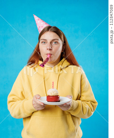 Young happy woman in birthday party hat with playing pipe holding cake on blue background. Emotional, young face. Human emotions, facial expression and holiday concept. 112847089