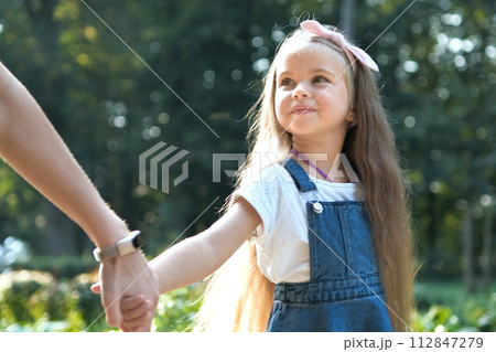 Young mom and her small daughter with long hair walking together holding hands in summer park. Young mom and her small daughter with long hair walking together holding hands in summer park. 112847279
