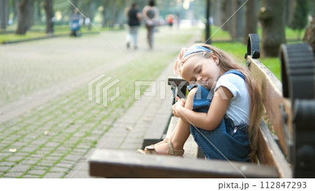 Small tired child girl sitting on a bench with closed eyes resting in summer park. 112847293