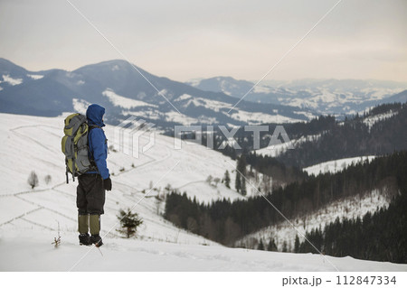 Back view of tourist hiker in warm clothing with backpack standing on mountain clearing on copy space background of woody mountain ridge and cloudy sky. Back view of tourist hiker in warm clothing with backpack standing on mountain clearing on copy space background of woody mountain ridge and cloudy sky. 112847334