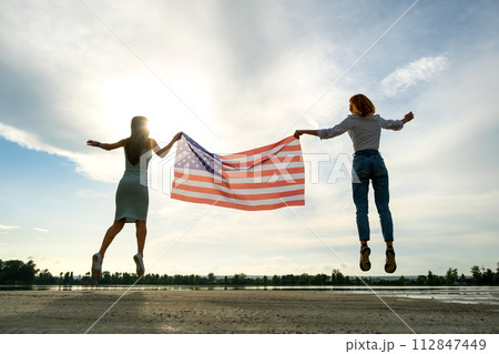Two young friends women holding USA national flag jumping up together outdoors at sunset. Patriotic girls celebrating United States independence day. Two young friends women holding USA national flag jumping up together outdoors at sunset. Patriotic girls celebrating United States independence day. 112847449