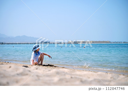 Lonely child girl playing with white sand on sea beach on background of blue sky, clear water and distant shore. Summer tourism and recreation concept Lonely child girl playing with white sand on sea beach on background of blue sky, clear water and distant shore. Summer tourism and recreation concept 112847746