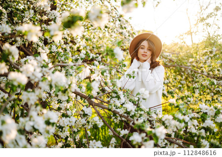 Portrait of Woman In hat posing near flowering tree. Smiling young woman enjoying smell of flowers. Portrait of Woman In hat posing near flowering tree. Smiling young woman enjoying smell of flowers. 112848616