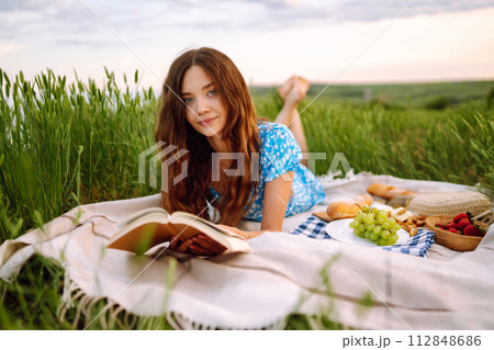 Young woman sits on a plaid with a book. Summer picnic in nature. Healthy food. 112848686