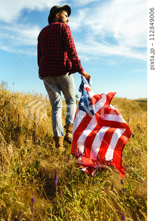 4th of July. American Flag. Traveler with the flag of America. The man in a hat, a backpack, a shirt and jeans. 4th of July. American Flag. Traveler with the flag of America. The man in a hat, a backpack, a shirt and jeans. 112849006