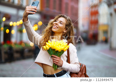 Selfie time. Young woman posing on the street with yellow flowers. Beautiful woman holding tulip. 112849475