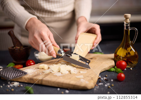 Woman slicing Piece of Traditional Italian Parmesan Hard cheese into flakes on a wooden cutting board at domestic kitchen 112855314