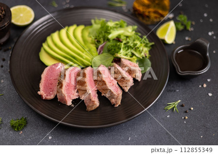 grilled cooked piece of tuna fillet with sliced avocado and salad on black ceramic plate 112855349