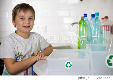 The child smiles, looking at the camera, ready to take apart the plastic. A child next to boxes with a recycling sign is ready to sort plastic. The concept of teaching a child to sort garbage. The child smiles, looking at the camera, ready to take apart the plastic. A child next to boxes with a recycling sign is ready to sort plastic. The concept of teaching a child to sort garbage. 112858577
