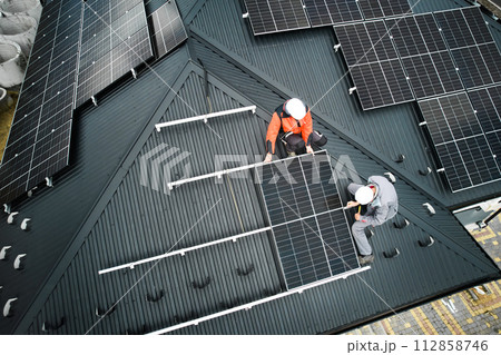 Mounters building photovoltaic solar module station on roof of house. Men electricians in helmets installing solar panel system outdoors. Concept of alternative and renewable energy. Aerial view. Mounters building photovoltaic solar module station on roof of house. Men electricians in helmets installing solar panel system outdoors. Concept of alternative and renewable energy. Aerial view. 112858746