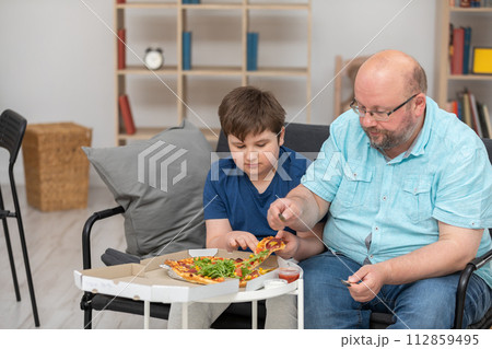 A father and son are sitting on the sofa sharing slices of pizza. A father and son are sitting on the sofa sharing slices of pizza. 112859495