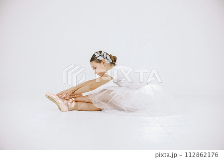 Little ballerina on white background. Brunette girl wearing white ballet suit. Girl sitting on a floor. Little ballerina on white background. Brunette girl wearing white ballet suit. Girl sitting on a floor. 112862176