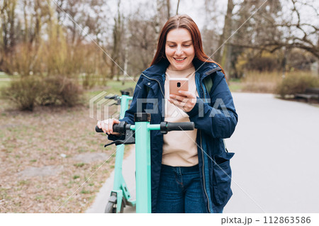 Smiling young woman standing near electric push scooter using smart phone. Ecological transport. Active lifestyle. Urban lifestyle concept. 112863586