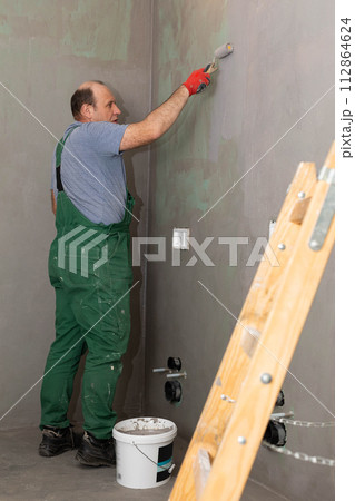 An experienced construction worker applies damp insulation with a paint roller to the wall. An experienced construction worker applies damp insulation with a paint roller to the wall. 112864624