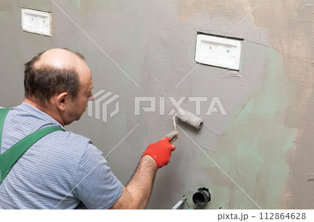 In the bathroom, a general construction worker applies moisture insulation using a roller. 112864628