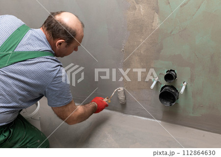 In the bathroom, a general construction worker applies moisture insulation using a roller. 112864630