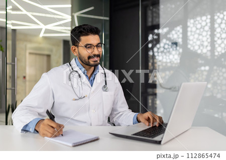 A doctor is sitting at a desk with a laptop and a notebook. He is writing something on the notebook A doctor is sitting at a desk with a laptop and a notebook. He is writing something on the notebook 112865474