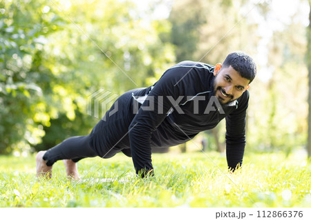 Active man in sportswear engaged in push-ups, showing strength and fitness at a sunny, green park. Active man in sportswear engaged in push-ups, showing strength and fitness at a sunny, green park. 112866376
