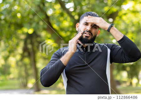 A tired male athlete wipes sweat from his forehead while touching his jaw, indicating pain, during an outdoor workout session. 112866486