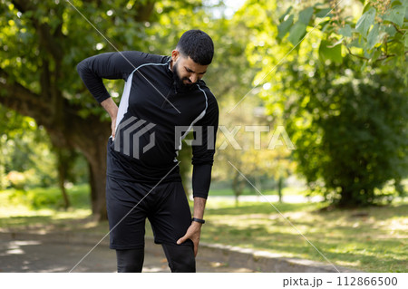 An athletic man in sportswear holds his lower back in pain while standing in a lush green park, showing signs of injury. 112866500
