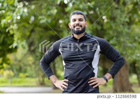 A young, fit man with a beard, smiling confidently while resting with his hands on his hips in a lush green park. A young, fit man with a beard, smiling confidently while resting with his hands on his hips in a lush green park. 112866598