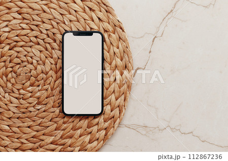 Smartphone with Blank screen on round rattan casket and beige marble background. Flat lay, top view minimalist, template or mockup. Smartphone with Blank screen on round rattan casket and beige marble background. Flat lay, top view minimalist, template or mockup. 112867236