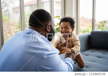 Portrait Of Happy African American Dad With Cute Little Baby Girl on couch at home in the living room, caring father smiling and amusing his girl while sitting on the couch, happy family Portrait Of Happy African American Dad With Cute Little Baby Girl on couch at home in the living room, caring father smiling and amusing his girl while sitting on the couch, happy family 112868130