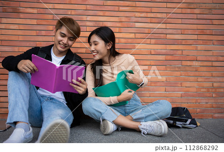 Group of young interracial diverse university students reading textbook and sitting outside a classroom under a building, engaging in a discussion together, college campus, enjoying campus recreation. 112868292