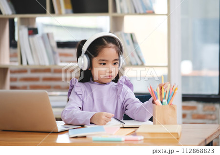 A young girl wearing headphones is sitting at a desk with a laptop 112868823