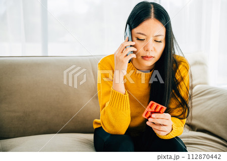Highlighting consultation, Asian woman takes medicine consults doctor via phone while on couch at home. Depicting healthcare quarantine and social distancing amid coronavirus. Highlighting consultation, Asian woman takes medicine consults doctor via phone while on couch at home. Depicting healthcare quarantine and social distancing amid coronavirus. 112870442