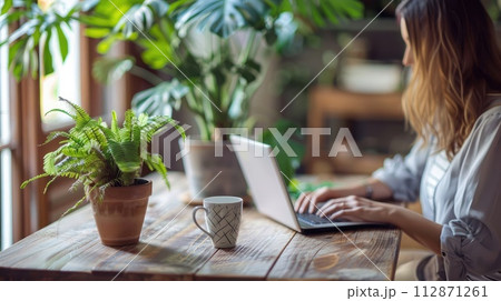 woman sitting at wooden table and working on laptop. A cup of coffee with a potted plant. The environment is bright and clean, a relaxed work environment 112871261