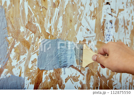 Interior painting, house painter applies brush strokes with gray paint to chipboard wall. 112875550